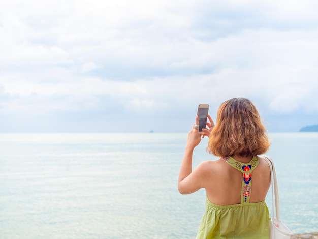 Hermosa mujer asiática con el pelo corto usando un teléfono inteligente toma una foto con vistas al paisaje marino