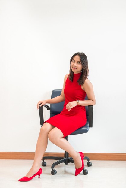 Hermosa mujer asiática en pose de vestido rojo para tomar una foto en la silla sobre fondo de pared blanca