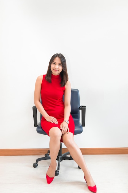 Hermosa mujer asiática en pose de vestido rojo para tomar una foto en la silla sobre fondo de pared blanca