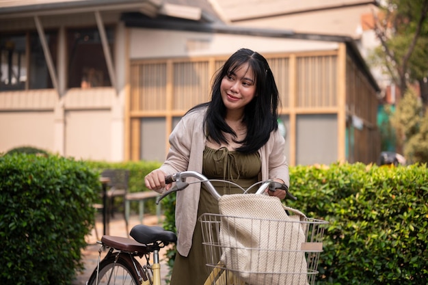 Una hermosa mujer asiática positiva en un vestido lindo está caminando y empujando su bicicleta en la ciudad