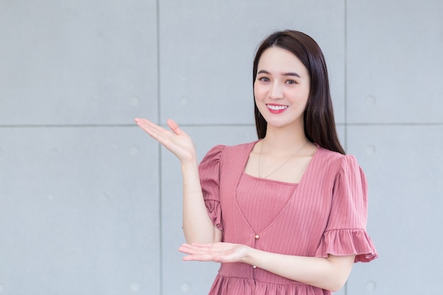 Hermosa mujer asiática que viste un vestido rosa está de pie y sonriendo muestra la mano para presentar algo