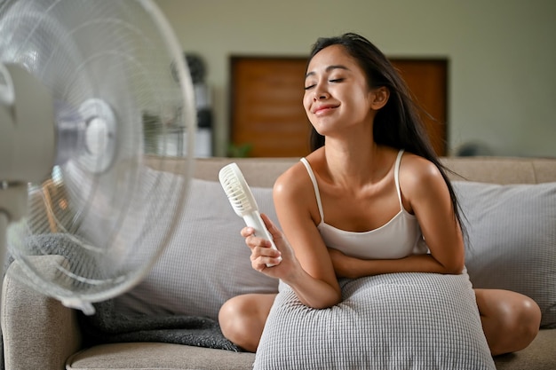 Una hermosa mujer asiática se refresca con un práctico ventilador y un ventilador mientras está sentada en un sofá
