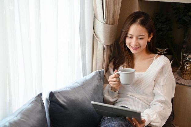 Hermosa mujer asiática relajándose en la sala de estar en el sofá tomando café caliente mientras usa la tableta