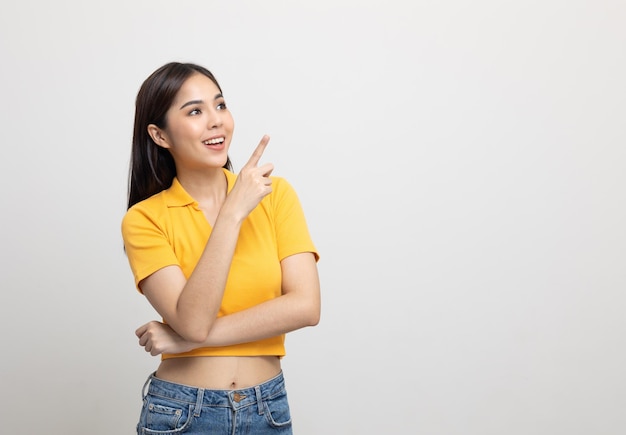 Hermosa mujer asiática señalando con el dedo al espacio en blanco. Mujer sonriente con camisa amarilla sobre fondo blanco aislado. Chica adolescente atractiva mirando el espacio para el texto.