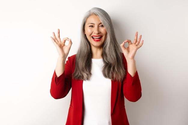 Hermosa mujer asiática senior en chaqueta roja, sonriendo y mostrando signos de aprobación, aprueba y dice que sí, de pie sobre fondo blanco.