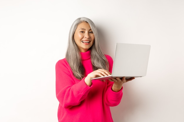 Hermosa mujer asiática senior empresaria trabajando con laptop riendo y sonriendo a la cámara de pie ...