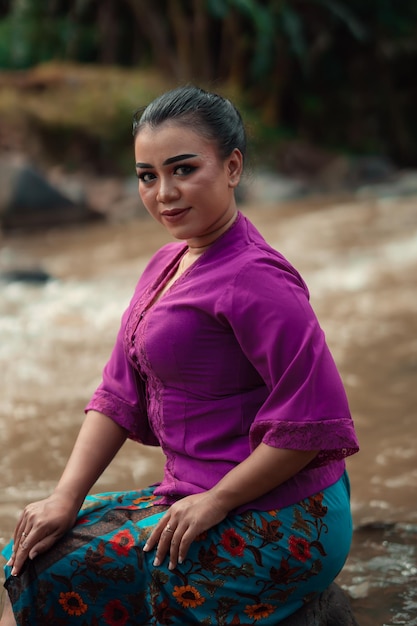 Hermosa mujer asiática sentada en la roca cerca del río con una cara sonriente mientras usa un vestido morado