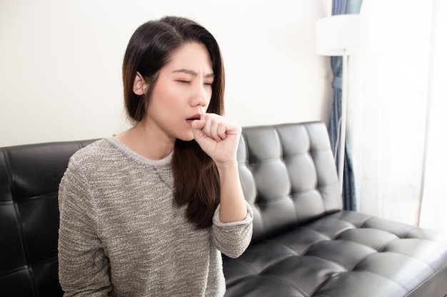 Hermosa mujer asiática sentada en la sala de estar tiene tos y con la mano toca su pecho Un concepto de salud