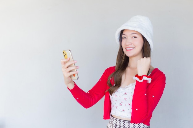 Hermosa mujer asiática smilinhowho usa abrigo rojo y sombrero blanco como una niña Santy sostiene un teléfono inteligente.