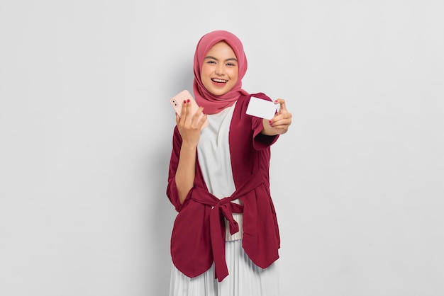 Hermosa mujer asiática sonriente con camisa casual e hiyab sosteniendo un teléfono móvil, mostrando tarjeta de crédito, regocijándose de la suerte aislada sobre fondo blanco. Concepto de estilo de vida religioso de personas