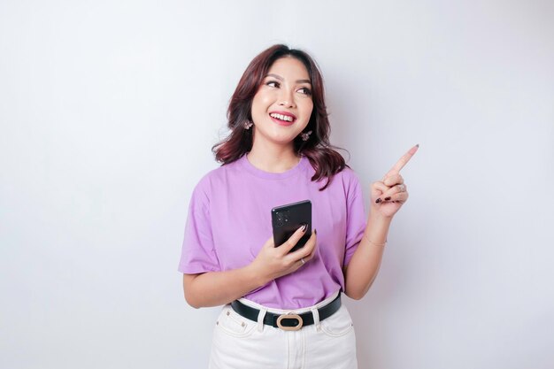 Hermosa mujer asiática sonriente señalando con el dedo el espacio vacío a su lado mientras sostenía su teléfono con fondo blanco aislado