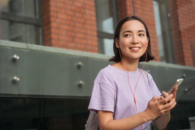 Hermosa mujer asiática sonriente sosteniendo un teléfono móvil escuchando música en la calle, copiando espacio