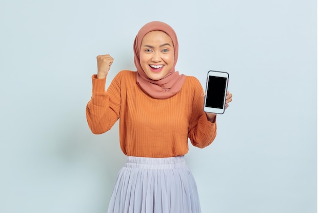 Hermosa mujer asiática sonriente con suéter marrón y hiyab mostrando la pantalla del teléfono móvil en blanco y mostrando un gesto celebrando la suerte aislada en el concepto de estilo de vida musulmán de fondo blanco