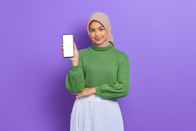 Hermosa mujer asiática sonriente con suéter verde y hiyab mostrando un teléfono móvil con pantalla en blanco aislado sobre un fondo morado