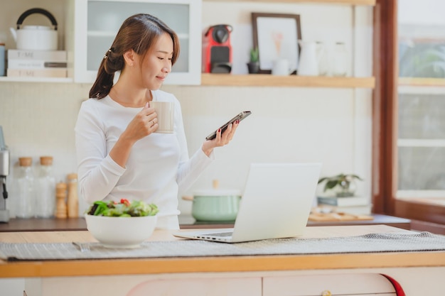 Hermosa mujer asiática sonriente usando un teléfono inteligente en la cocina mientras bebe café por la mañana