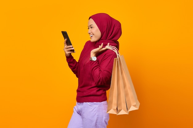 Hermosa mujer asiática sosteniendo bolsas de la compra y con smartphone sobre fondo amarillo