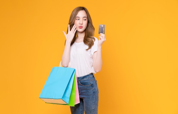 Hermosa mujer asiática sosteniendo bolsas de papel marrón en blanco y mostrando tarjeta de crédito con fondo amarillo