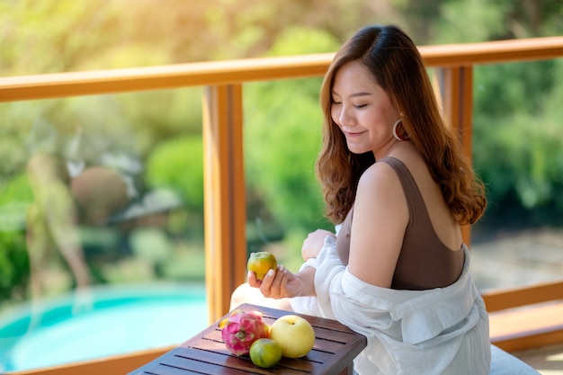 Una hermosa mujer asiática sosteniendo frutas mientras disfruta sentado en el balcón con el fondo de vista al mar