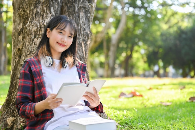 Hermosa mujer asiática sosteniendo un libro sonriendo y mirando a la cámara mientras se sienta bajo el árbol