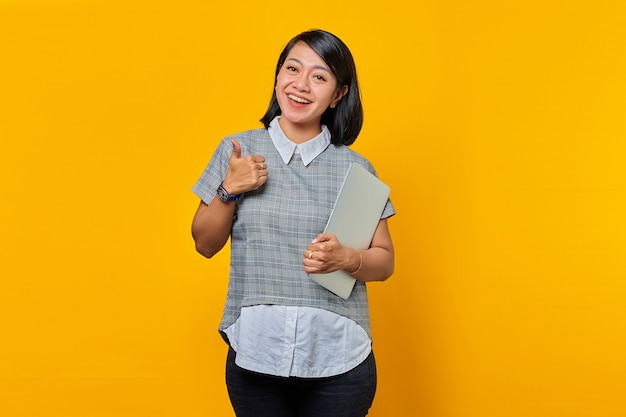 Hermosa mujer asiática sosteniendo portátil sonriendo y mostrando Thumbs up sign sobre fondo amarillo