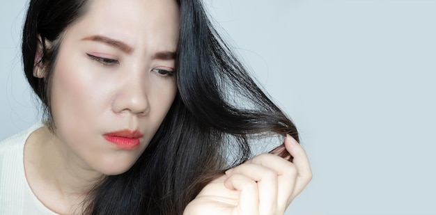 Hermosa mujer asiática sostiene y mira su cabello de punta para ver el cabello dividido y seco con cara de preocupación