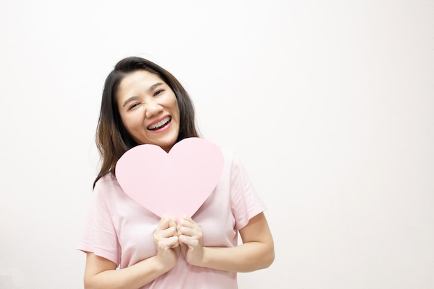 Hermosa mujer asiática sostiene papel con forma de corazón rosa frente a sí misma con una sonrisa y felicidad. un dia de san valentin y amor