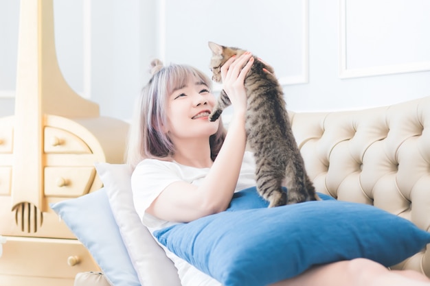 Una hermosa mujer asiática, tailandesa, está acostada con un gato en el sofá con una cara sonriente y feliz en la sala de estar