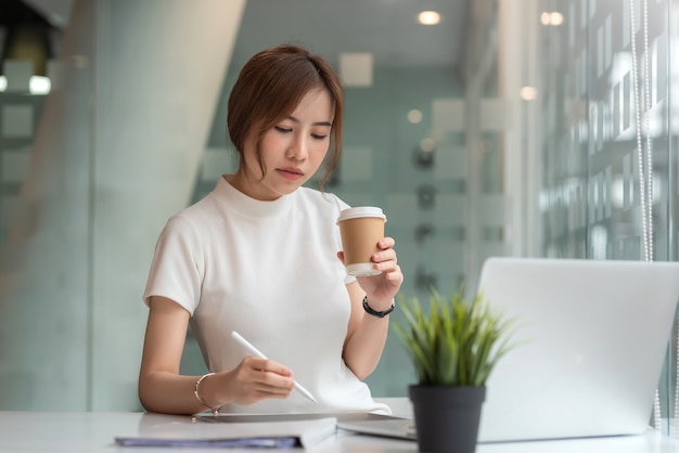 Hermosa mujer asiática con taza de café sentado trabajando portátil en la oficina moderna.