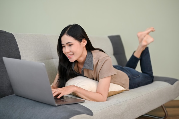 Hermosa mujer asiática tendida en un sofá en la sala de estar usando una laptop para chatear con sus amigos