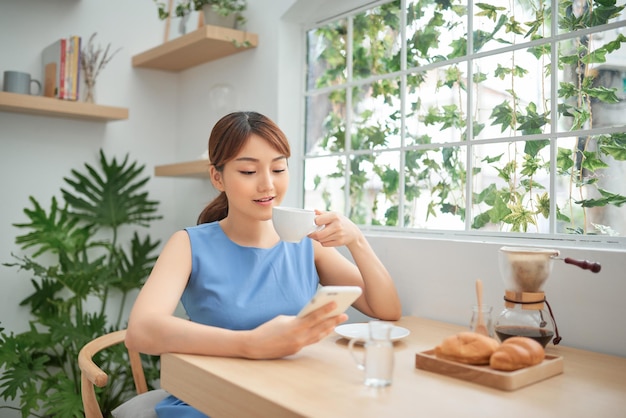 Hermosa mujer asiática tomando café y usando el teléfono detrás de la ventana