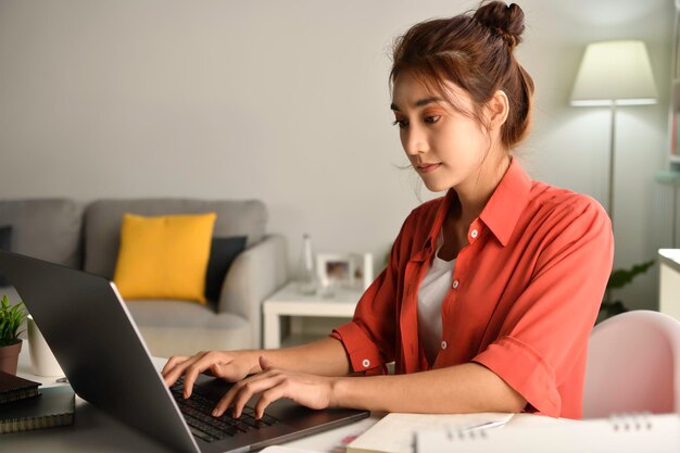 Hermosa mujer asiática trabajando en una laptop en casa. Estudiante sentada en el escritorio, estudio en línea en la computadora portátil, tome notas en el cuaderno