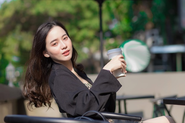 Hermosa mujer asiática con traje negro está sentada en una silla y sonriendo frente a la cafetería