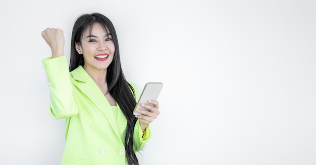 Hermosa mujer asiática en traje verde pastel usando teléfono inteligente gesticulando sí aislado sobre fondo blanco Feliz chica alegre con cabello largo celebrando la victoria mujer asiática exitosa en las redes sociales