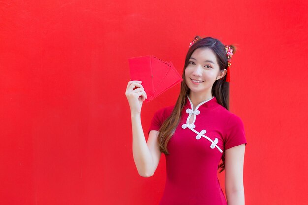 Hermosa mujer asiática usa un cheongsam rojo y sostiene sobres rojos mientras sonríe con fondo rojo