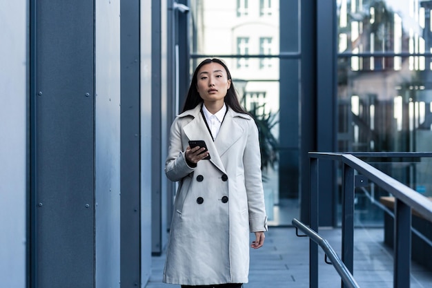 Hermosa mujer asiática vestida de negocios usa el teléfono, camina cerca del moderno centro de oficinas afuera