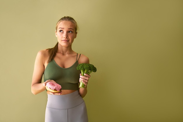 Hermosa mujer atlética y deportiva parada cerca de un fondo verde sosteniendo una rosquilla rosa en una mano y brócoli en la otra Dudas sobre la elección de alimentos poco saludables y saludables