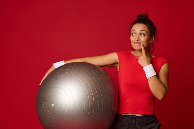 Hermosa mujer atlética multiétnica pensativa sosteniendo una pelota en forma y mirando cuidadosamente a un lado aislada sobre fondo de color rojo con espacio de copia