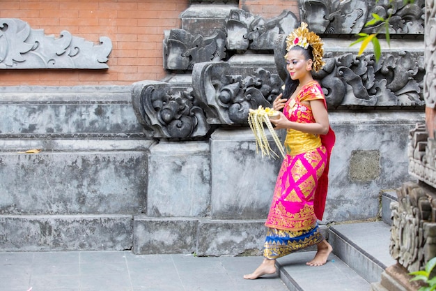 Foto hermosa mujer balinesa con ropa tradicional y caminando en un templo mientras lleva un cuenco de ofrendas