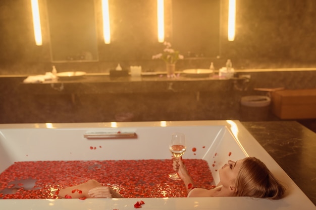 Hermosa mujer en baño con pétalos de rosa bebiendo vino