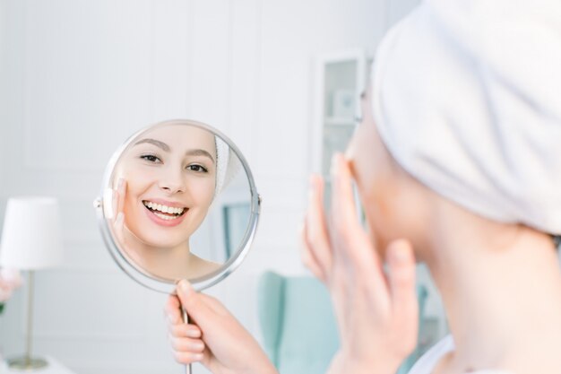 Hermosa mujer en bata de baño blanca y toalla mirando su piel perfecta en el espejo y aplicando la base de crema tonal en la cara