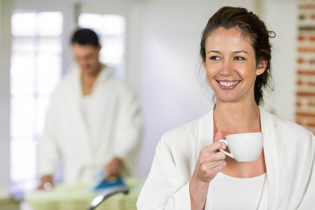 Hermosa mujer en bata de baño con una taza de té en la cocina y el hombre planchando la ropa detrás de ella