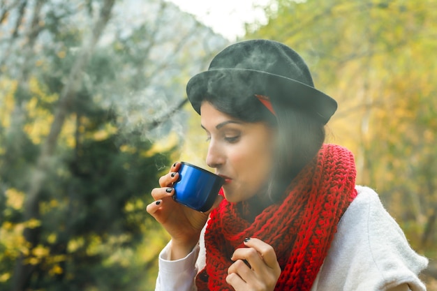 Hermosa mujer en una bata blanca crochet bufanda roja y sombrero negro tiene una taza de té en sus manos