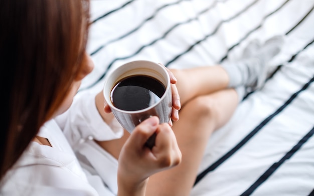 Una hermosa mujer bebiendo café caliente en una cama blanca y acogedora en su casa por la mañana