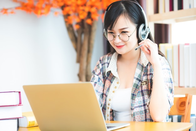 Hermosa mujer blanca asiática con gafas y auriculares está felizmente usando una computadora portátil para estudiar en línea.