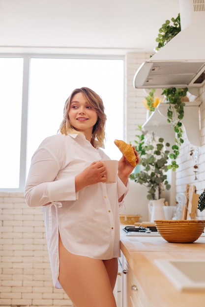 Hermosa mujer bonita de pie en la cocina mientras toma un café con un croissant