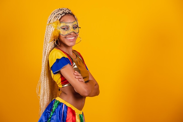 Hermosa mujer brasileña negra vestida con ropa de carnaval máscara fondo amarillo brazos cruzados