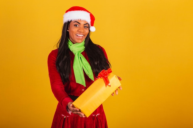 Hermosa mujer brasileña negra vestida con ropa de Navidad Santa Claus entregando un regalo amarillo