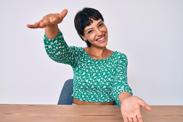 Hermosa mujer brunettte con ropa informal sentada en la mesa mirando a la cámara sonriendo con los brazos abiertos para abrazar la expresión alegre abrazando la felicidad