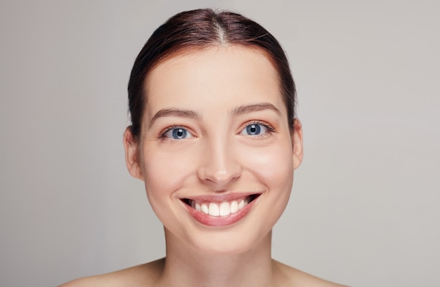 Hermosa mujer con cabello castaño, piel limpia y fresca posando en un estudio gris, mirando lacio, sonriendo ampliamente, modelo con ligero maquillaje desnudo