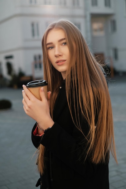 Hermosa mujer con cabello largo con café para llevar en la calle Estudiante femenina paseando Coffee break